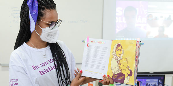 Imagem mostra uma voluntária que está segurando um livro com a ilustração da ativista Malala.