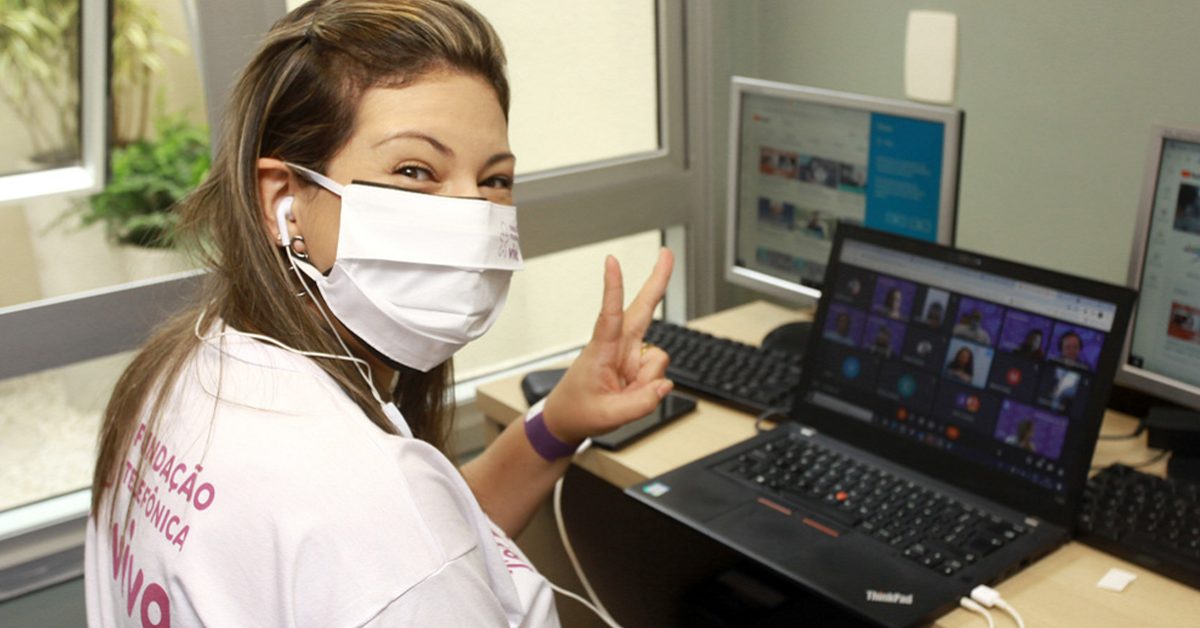 Imagem mostra voluntária. Ela usa máscara, está sorrindo e sentada em frente a um notebook assistindo a uma live do Dia dos Voluntários