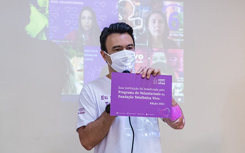 Imagem mostra voluntário com uma placa com informações sobre uma instituição beneficiada no Dia dos Voluntários