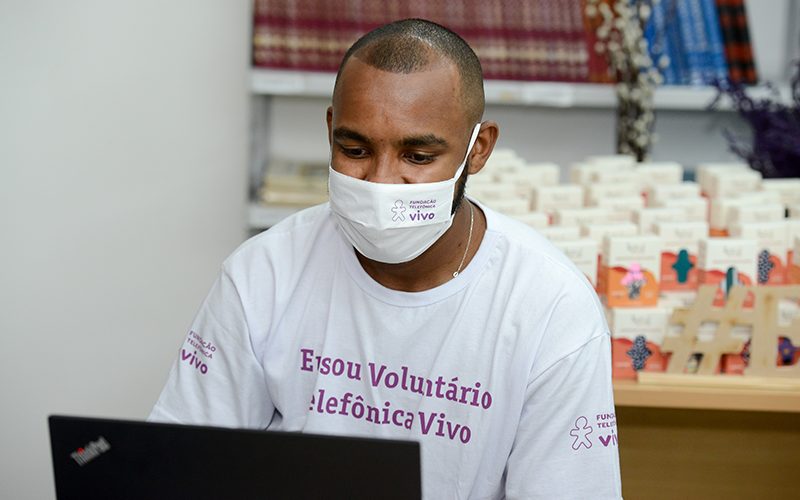 Imagem mostra um voluntário. Ele usa máscara e veste a camiseta do Dia dos voluntários. Ele está usando um notebook.