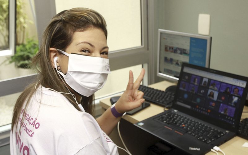 Imagem mostra voluntária. Ela usa máscara, está sorrindo e sentada em frente a um notebook assistindo a uma live do Dia dos Voluntários