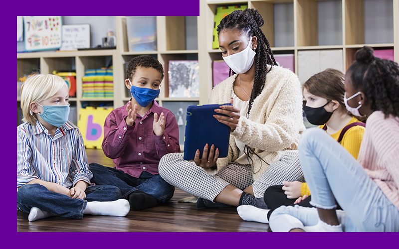 Imagem mostra professora com quatro alunos. Todos usam máscara e estão sentados no chão de uma sala de aula. A professora segura um tablet e está mostrando o aparelho para um dos alunos.