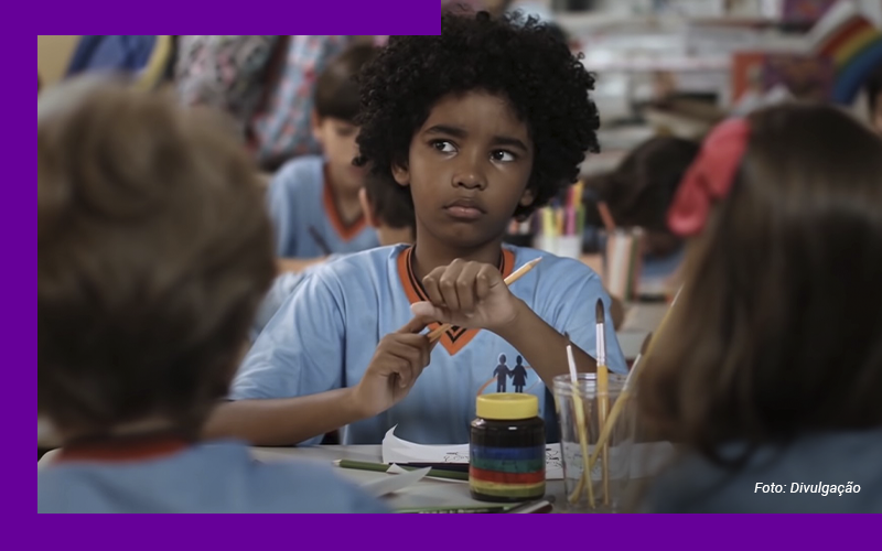 Imagem mostra um menino negro, com uniforme escolar, sentado em uma mesa com outras crianças. Ele segura um lápis de cor. Sobre a mesa, há outros lápis, pincéis, papel e um frasco de tinta