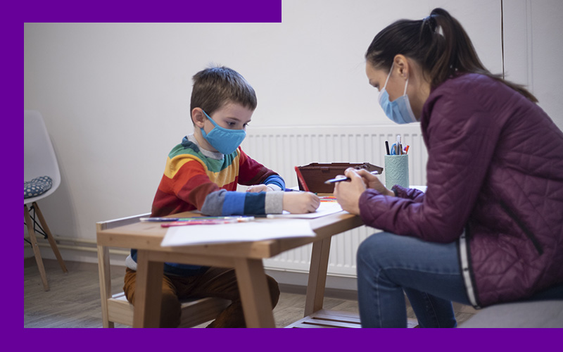 Imagem mostra uma psicopedagoga auxiliando uma criança em sala de aula. A criança está sentado em uma cadeira e em uma mesa há objetos pedagógicos. A profissional está em sua frente.