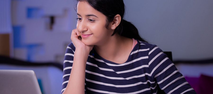 Imagem mostra uma jovem olhando para a tela de um computador. Ela usa blusa azul marinho listrada, tem cabelos compridos, que estão presos, e está sorrindo.