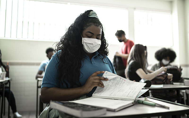 Imagem mostra uma estudante em sala de aula. Ela tem cabelos longos, que está preso, usa máscara e uniforme na cor azul. Está manuseando um caderno e está sentada em uma carteira escolar. Ao fundo, outros alunos aparecem na imagem.