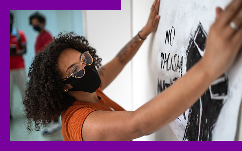 Imagem mostra uma jovem negra fixando em uma parede um cartaz contra o racismo