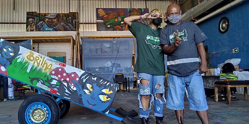 Imagem mostra dois catadores de materiais recicláveis em um galpão do projeto Pimp my carroça. Eles estão em frente a uma carroça personalizada com um grafite.