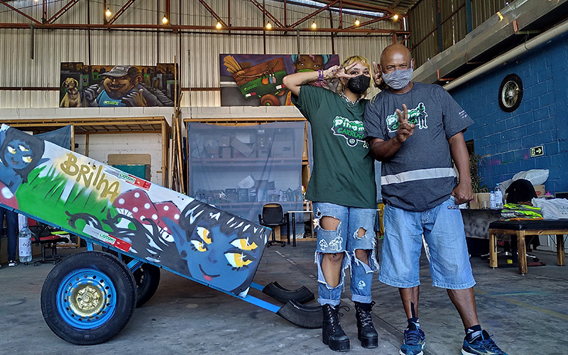 Imagem mostra dois catadores de materiais recicláveis em um galpão do projeto Pimp my carroça. Eles estão em frente a uma carroça personalizada com um grafite.