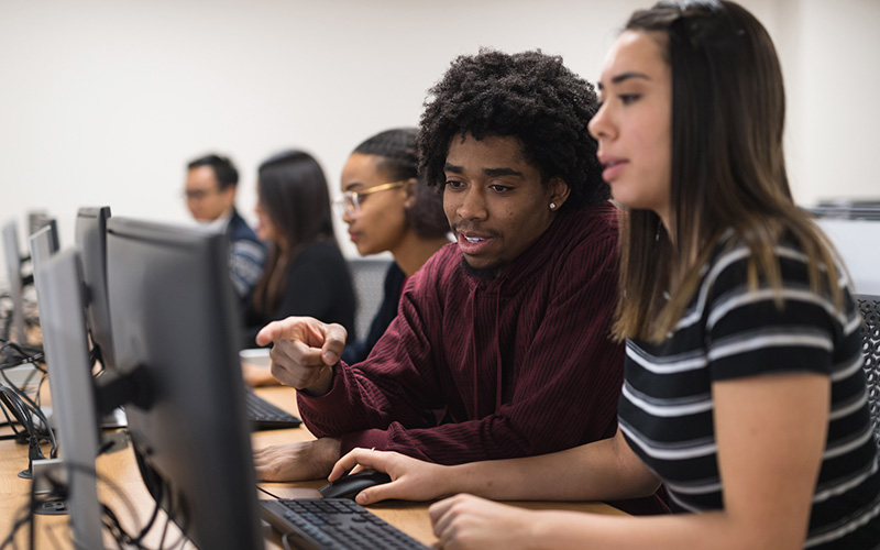 Imagem mostra cinco estudantes em uma sala com computadores. Em primeiro plano, é possível ver um estudante e uma estudante conversando enquanto olham para a tela do computador. O jovem é negro e usa uma blusa na cor vinho e a jovem é branca e uma camiseta listrada.