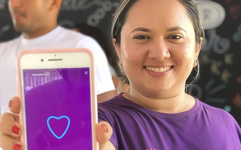 Imagem mostra a voluntária Geciclelma. Ela usa uma camiseta na cor roxa, está segurando um celular com a mão direita e mostrando ele para a câmera. Ela está sorrindo. Na tela do celular, há uma imagem na cor roxa com a ilustração de um coração.