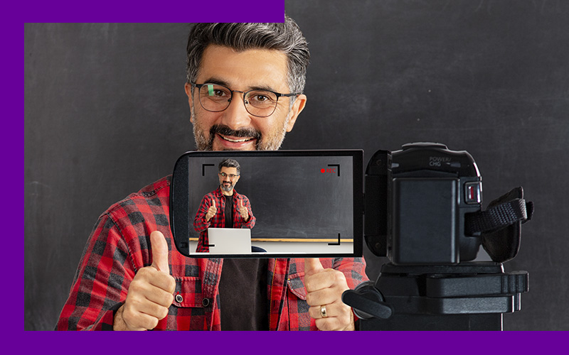 Imagem mostra um educador em frente a uma câmara de filmagem. Ele usa uma camisa xadrez e óculos. Está sorrindo e fazendo sinal de “positivo” para a câmera. Ao fundo, é possível ver uma lousa, o ambiente parece ser uma sala de aula.
