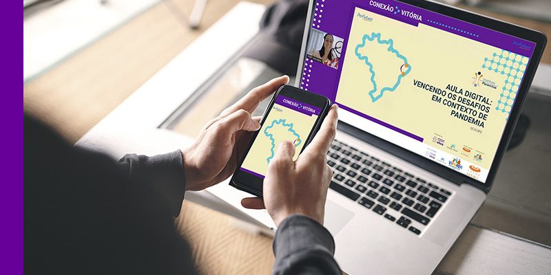Imagem mostra um computador ligado em que é possível ver a tela inicial do evento Conexão Vitória, com a participação de uma educadora. Também é possível ver a mão de uma pessoa que parece estar assistindo ao evento pelo computador e está segurando um smartphone em que é possível ver o mapa do Brasil na tela