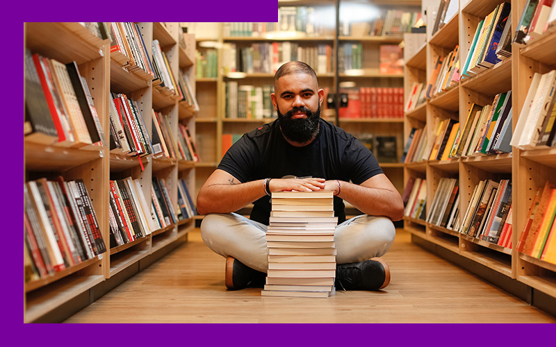 O empreendedor social Tiago Silva está sentando no chão de uma biblioteca, com as pernas cruzadas. A sua frente uma pilha de livros, na qual ele está com as mãos apoiadas.