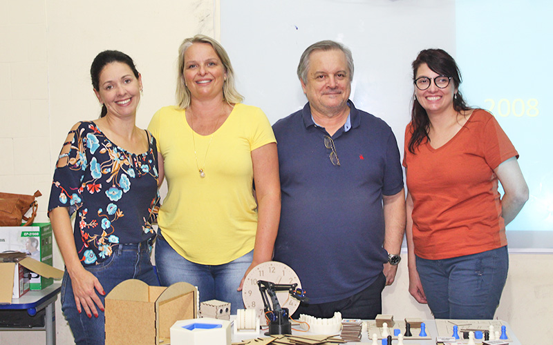 Imagem mostra as três professoras e um professor que fazem parte da equipe responsável pelo projeto vencedor do prêmio enlighted. Eles estão lado a lado em um ambiente que parece ser um laboratório. É possível ver uma mesa e alguns objetos pedagógicos em cima dela.