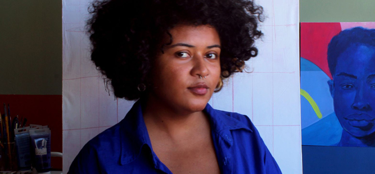 Imagem mostra a jovem Kika Carvalho. Ela é negra e tem os cabelos ondulados e curtos. Está usando uma camisa azul. Ao fundo, é possível ver uma de suas obras