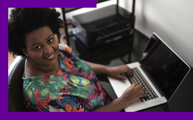 Professora usa um laptop e sorri para a foto. Ela é uma mulher negra e usa uma blusa verde com estampa de flores