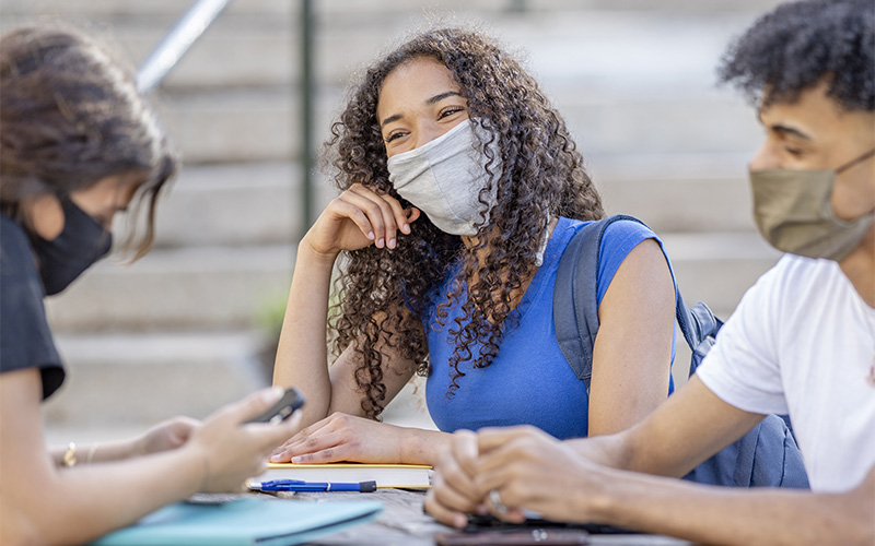 três jovens estudantes, com máscaras de proteção, conversam em volta de uma mesa.