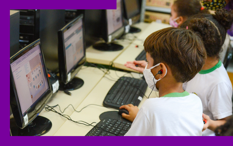 Imagem mostra dois alunos utilizando computadores. Eles estão usando uniforme escolar e máscara.