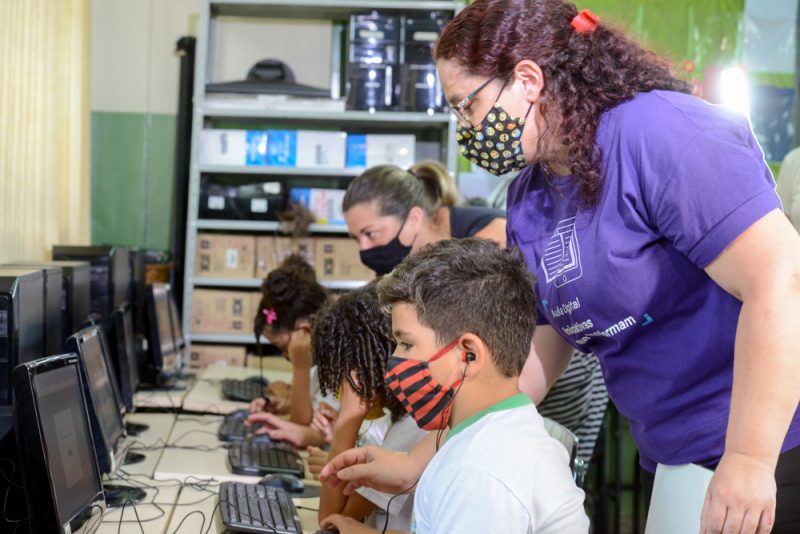 Imagem mostra duas educadoras auxiliando os alunos que estão utilizando computadores em sala de aula.