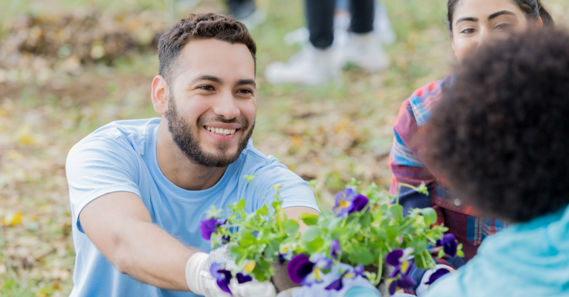 Imagem mostra três jovens em um local que parece ser um parque. Um dos jovens está segurando um vaso de flores e entregando para uma das jovens. A terceira jovem observa os dois jovens interagindo.