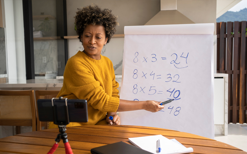 Imagem mostra uma educadora ensinando matemática. Ela está apontando para alguns números apresentados em um painel. À sua frente há um celular, o que nos faz entender que a aula que ela está dando é on-line.