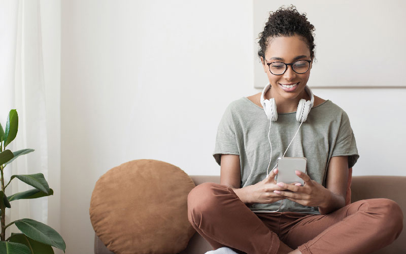 Mulher negra sentada sobre o sofá, com fone de ouvidos pendurados ao pescoço, interagindo com smartphone à mão.