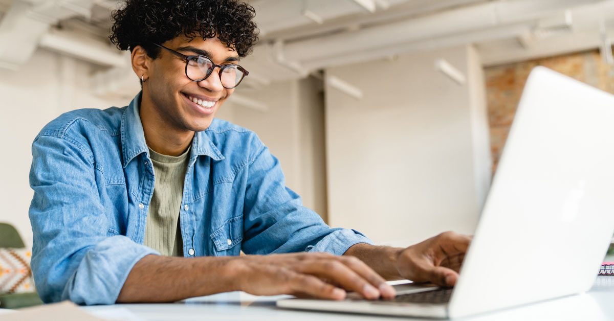 Imagem mostra um jovem utilizando um notebook. Ele é negro, usa óculos, camisa jeans e está sorrindo.
