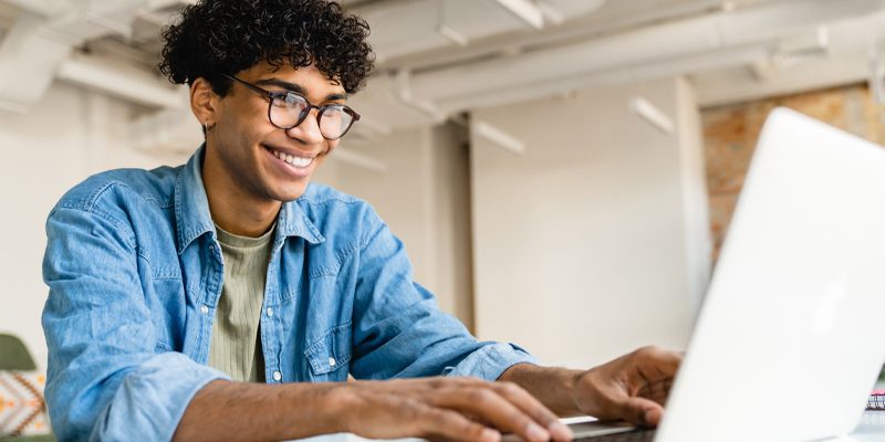 Imagem mostra um jovem utilizando um notebook. Ele é negro, usa óculos, camisa jeans e está sorrindo.