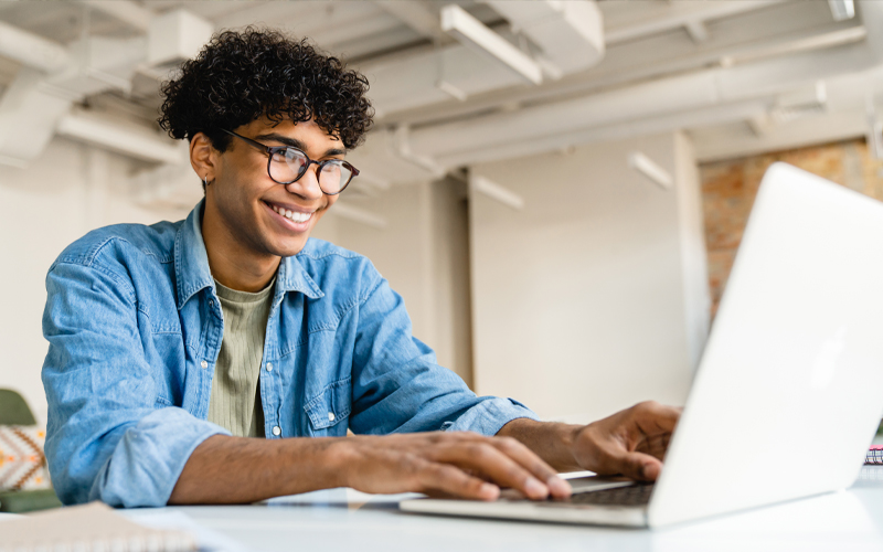 Imagem mostra um jovem utilizando um notebook. Ele é negro, usa óculos, camisa jeans e está sorrindo.