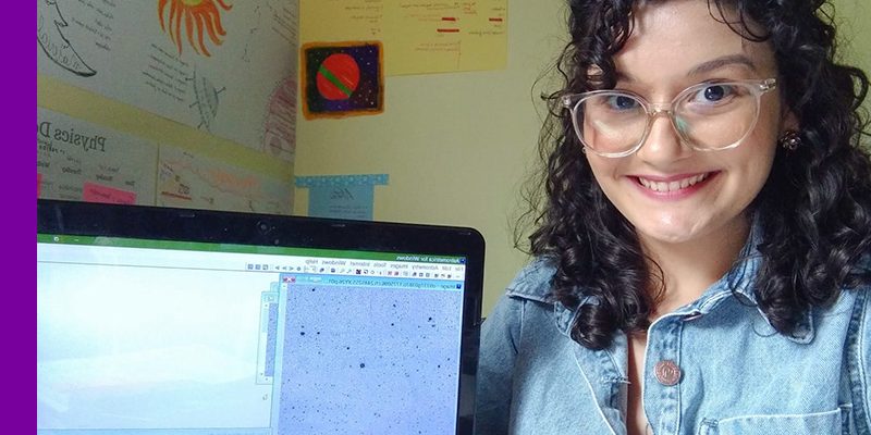 Imagem mostra a jovem Geovana Ramos. Ela está sorrindo, usa óculos, uma camisa jeans e tem os cabelos cacheados. Ao seu lado, há um computador em que é possível ver na tela uma possível imagem do espaço. Ao fundo, há imagens fixadas na parede do ambiente.