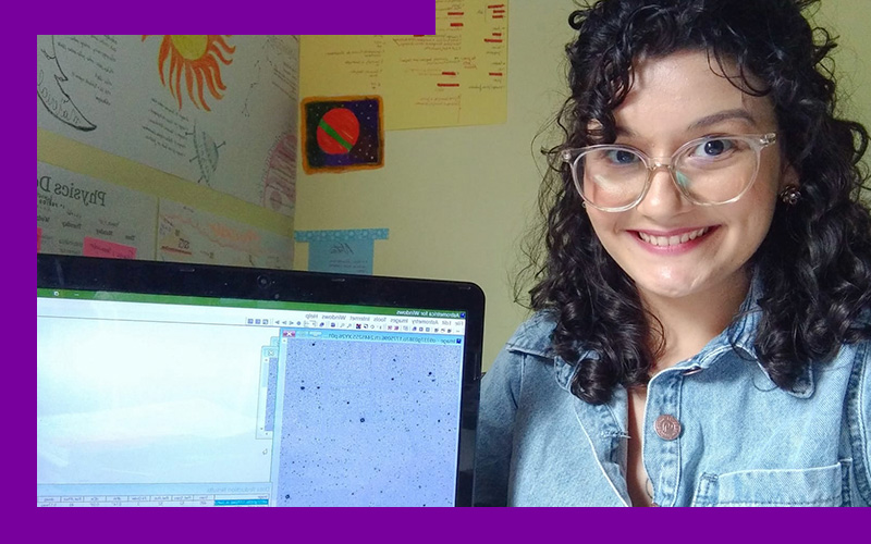 Imagem mostra a jovem Geovana Ramos. Ela está sorrindo, usa óculos, uma camisa jeans e tem os cabelos cacheados. Ao seu lado, há um computador em que é possível ver na tela uma possível imagem do espaço. Ao fundo, há imagens fixadas na parede do ambiente.
