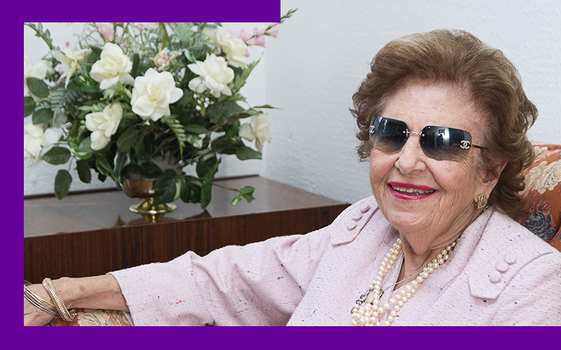 Imagem mostra Dorina Nowill. Ela é uma senhora idosa, está usando óculos escuros e uma camisa na cor rosa claro. Ao fundo, é possível ver uma mesa com um vaso de flores brancas em cima.