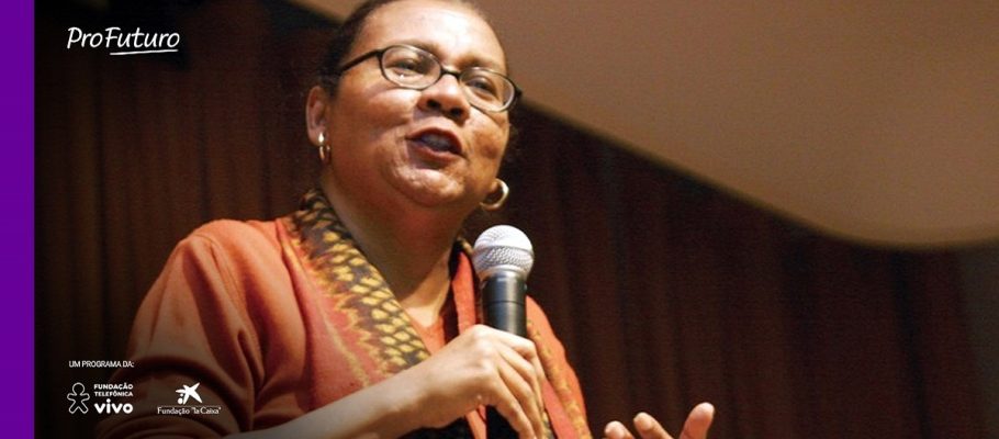 Imagem mostra a ativista bell hooks. Ela está segurando um microfone e parece estar palestrando. Usa óculos, brincos dourados e uma blusa vermelha.