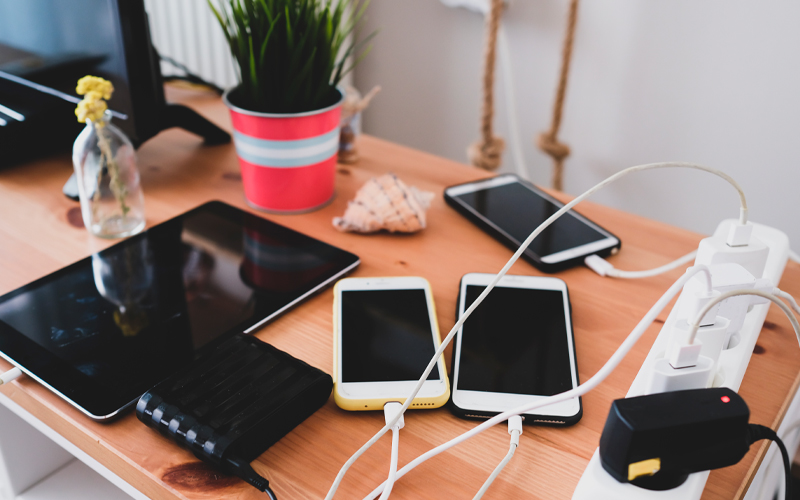 Imagem mostra uma mesa com diversos aparelhos eletrônicos como celulares, tablet e a o monitor de um computador. Os três aparelhos de celular estão conectados a cabos e estão sendo carregados em tomadas