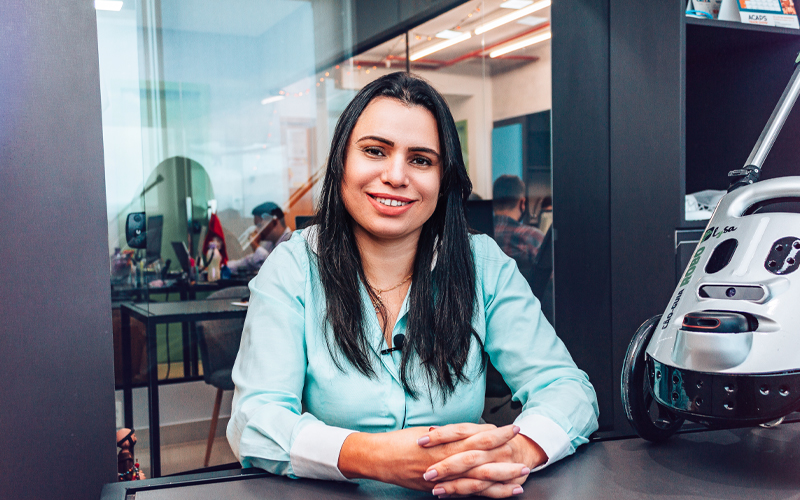 Imagem mostra a professora Neide Sellin, criadora do cão-guia robô. Ela usa uma camisa azul, tem cabelo comprido, está sorrindo para a foto e com os braços apoiados em uma mesa. Ao seu lado, é possível ver o robô. Ao fundo, é possível ver algumas pessoas trabalhando em um ambiente que parece ser um escritório.