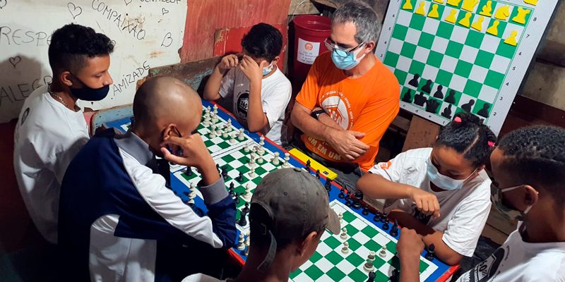 Imagem mostra alunos do projeto Casa Xadrez em volta de uma mesa onde há três tabuleiros do jogo. Eles estão acompanhados de um adulto, que parece ser o professor, e estão concentrados observando as peças do jogo