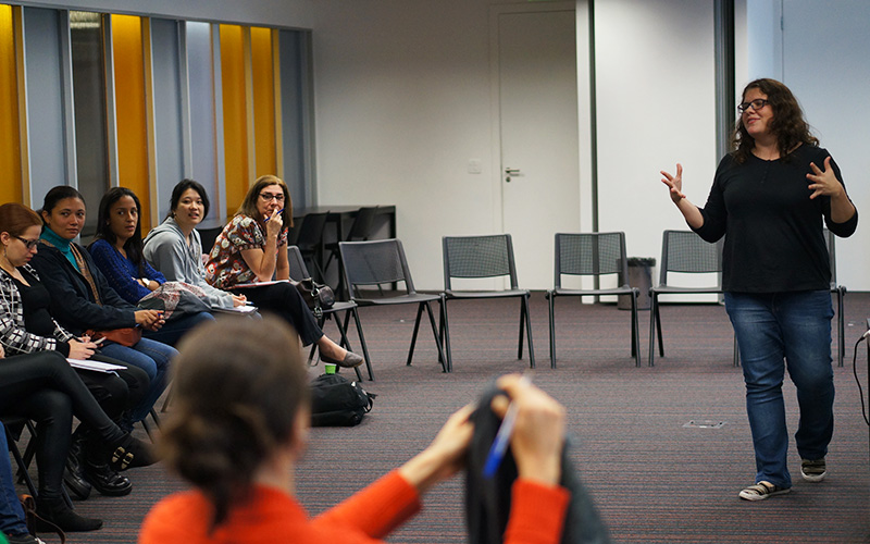 Imagem mostra uma sala de aula onde é possível ver alunas sentadas assistindo à aula de uma professora, que está em pé, conversando com as alunas.