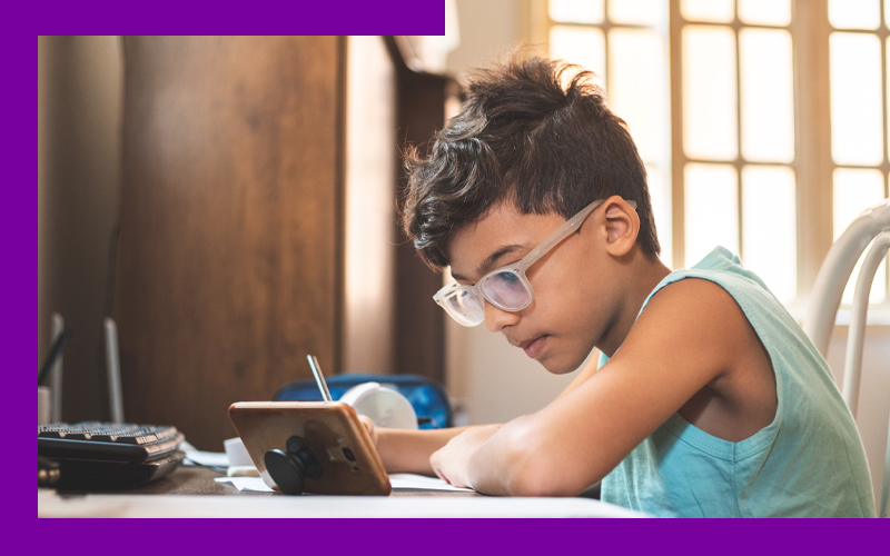 Imagem mostra um menino estudando com o apoio de um celular. Ele usa óculos, está com um lápis na mão direita, escrevendo em um caderno. O celular e o caderno estão sobre uma mesa.