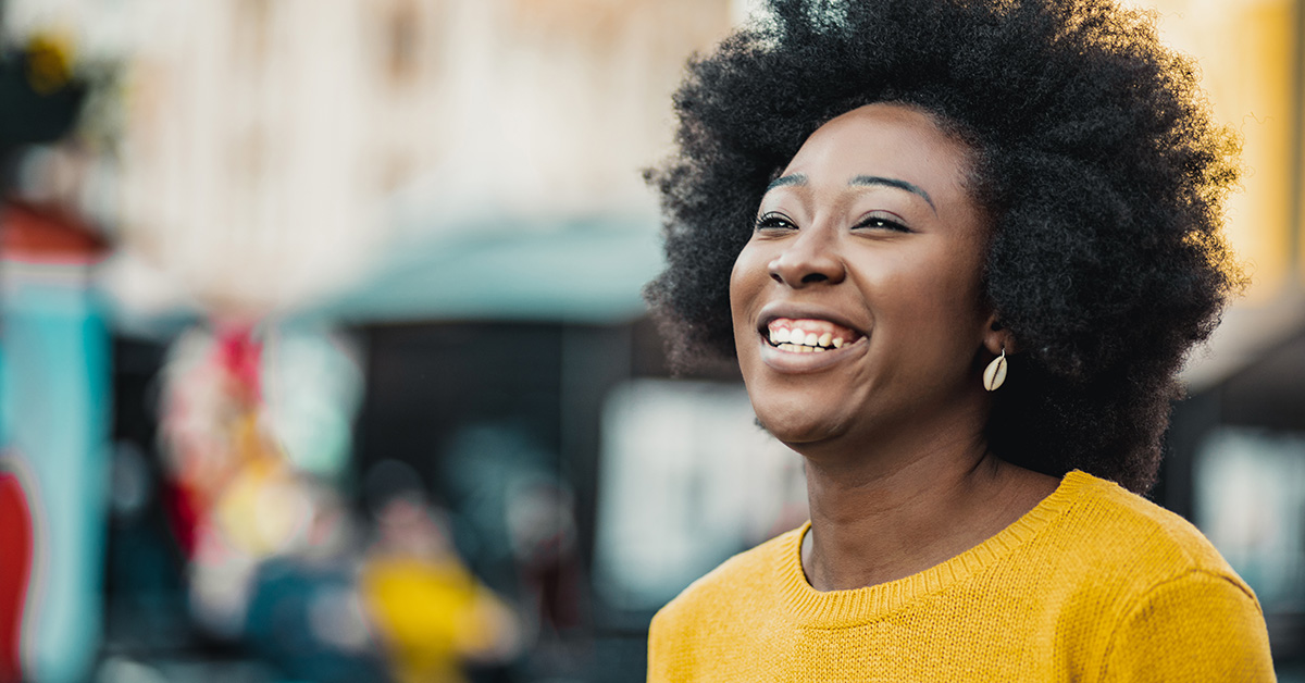 Imagem de mulher sorrindo representa live que tratou de temas como empreendedorismo na periferia e ancestralidade. Ela é negra, tem os cabelos crespos e cacheados e está usando uma blusa amarela