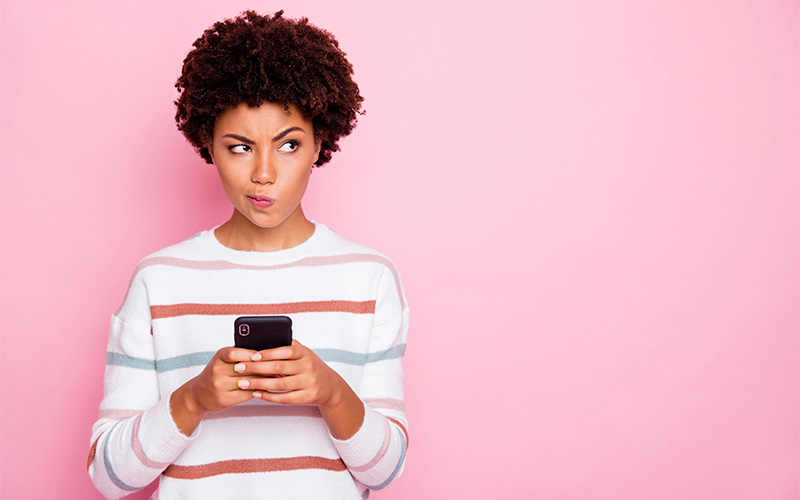 Imagem mostra uma jovem negra segurando um celular. Ela está com um semblante de dúvida. Usa blusa branca com listras nas cores azul e rosa.