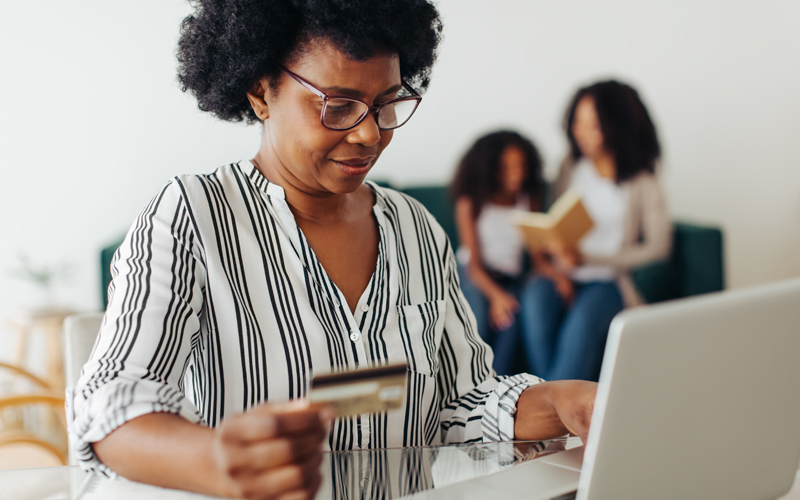 Imagem mostra uma mulher negra utilizando um notebook. Ela segura um cartão de crédito na mão direita, o que dá a entender que ela está fazendo uma transação financeira pela internet. Ao fundo, é possível ver uma mulher e uma menina sentadas em um sofá.