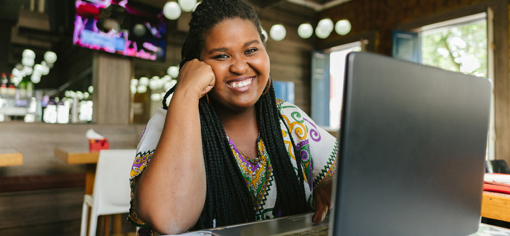 Imagem mostra uma jovem negra sorrindo. Ela usa tranças no cabelo, está sentada em frente a um notebook em um ambiente com outras mesas e uma televisão ligada ao fundo"
