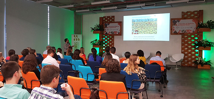 Mulher palestra do 1º Encontro de Negócios de Impacto Social. Outras quatro pessoas estão no palco, todas sentadas em cadeiras.
