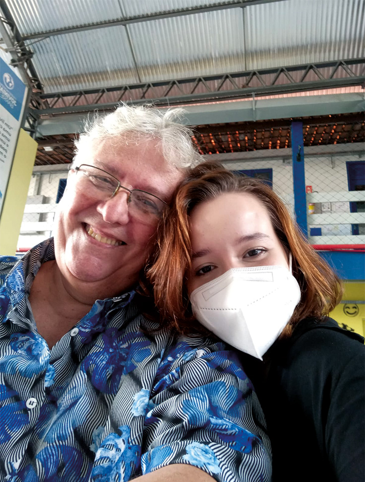 O professor Antero reencontra estudante Raquel Aquino para a entrega da carta. Os dois estão sorrindo, com os rostos próximos. Raquel é branca, tem os cabelos vermelhos e está de blusa preta e uma máscara de proteção individual. Ele é um senhor branco, de cabelos grisalhos e veste uma camisa de listras finas preta e branca com detalhes de flores azuis.