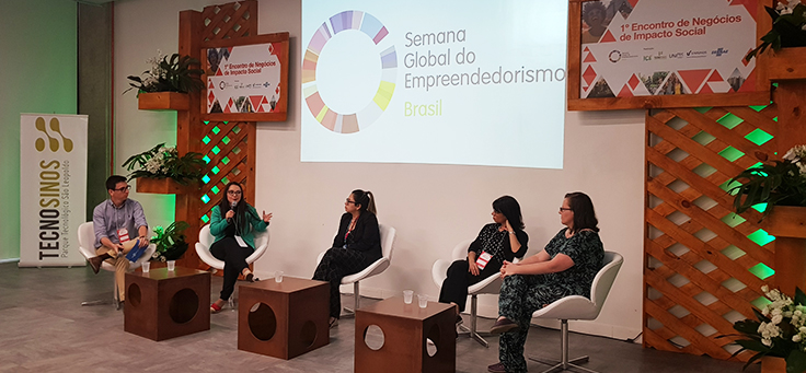Mulher palestra do 1º Encontro de Negócios de Impacto Social. Outras quatro pessoas estão no palco, todas sentadas em cadeiras.