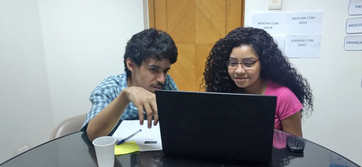 A jovem Karen Franquini e outro jovem rapaz, estão sentados em volta de uma mesa olhando para um notebook.