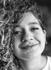 Caroline Monteiro Araújo, jornalista que montou o episódio sobre o Bellatucci Café olha para a câmera. Ela tem cabelos cacheados e está sorrindo.
