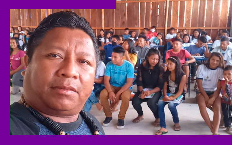 Imagem mostra o educador Juvêncio Cardoso. Ele está fazendo uma selfie e ao fundo é possível ver diversos alunos sentados em sala de aula.