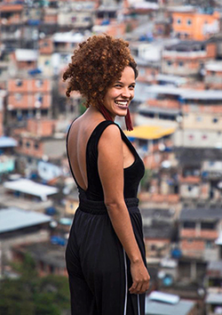 Thamyra Tamara, fundadora da GatoMídia que registrou a reação de jovens a vídeos 360° no melhor estilo Mecnologia, está sorrindo para foto, com os cabelos encaracolados presos e com o Complexo do Alemão ao fundo.
